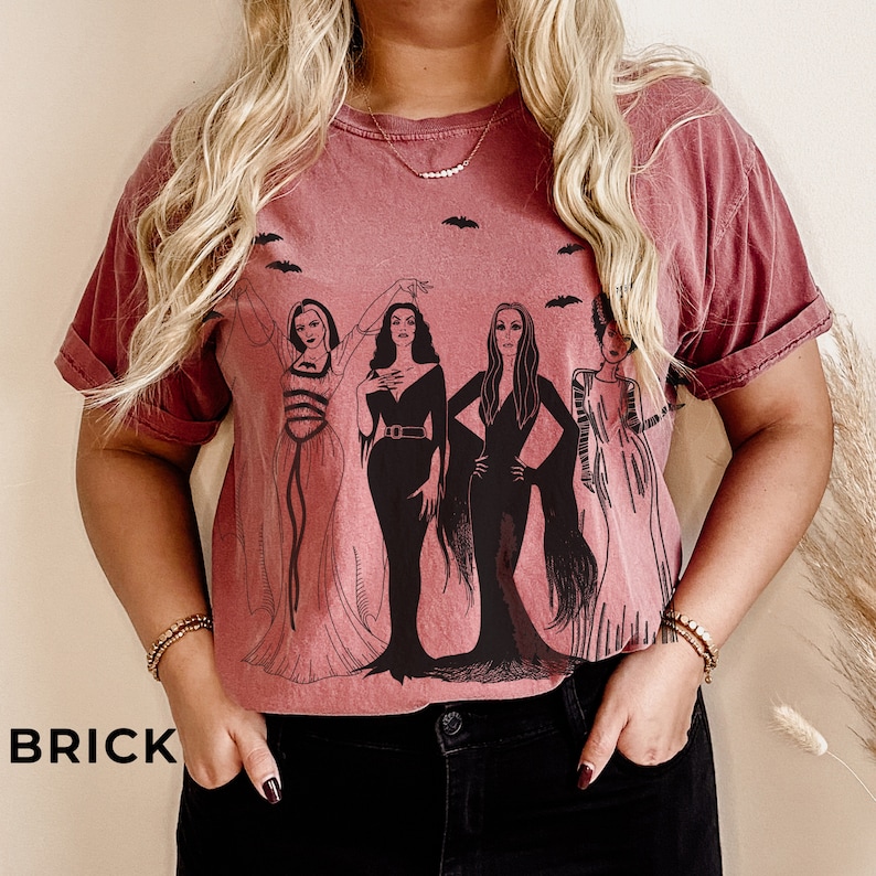 K&ouml;nnte beinhalten: Ein maronfarbenes T-Shirt mit einem grafischen Druck von vier Frauen in schwarzen Kleidern vor wei&szlig;em Hintergrund. Die Frauen tragen schwarze Kleider mit wei&szlig;en Akzenten und haben langes schwarzes Haar. Das Shirt hat einen Rundhalsausschnitt und kurze &Auml;rmel.