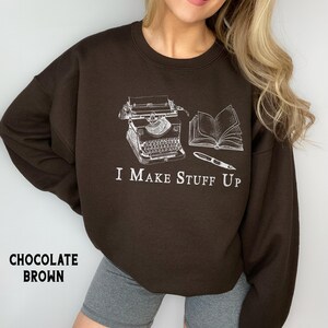 Puede incluir: Sudadera marr&oacute;n chocolate con un gr&aacute;fico blanco de una m&aacute;quina de escribir, un libro abierto y un bol&iacute;grafo. El texto "I Make Stuff Up" est&aacute; debajo. La sudadera es de cuello redondo.