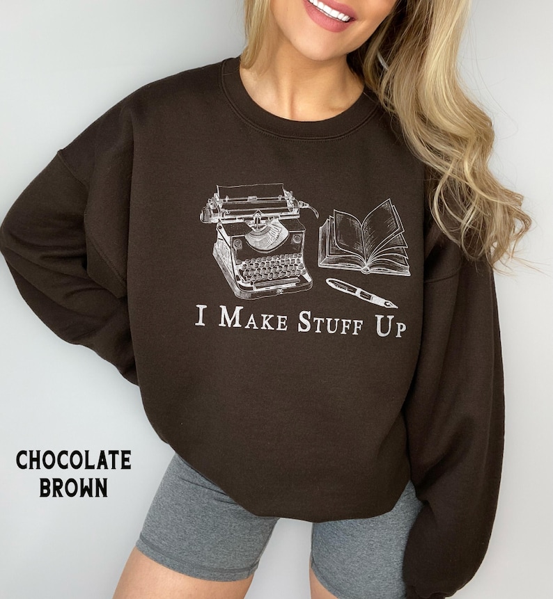 Puede incluir: Sudadera marr&oacute;n chocolate con un gr&aacute;fico blanco de una m&aacute;quina de escribir, un libro abierto y un bol&iacute;grafo. El texto "I Make Stuff Up" est&aacute; debajo. La sudadera es de cuello redondo.