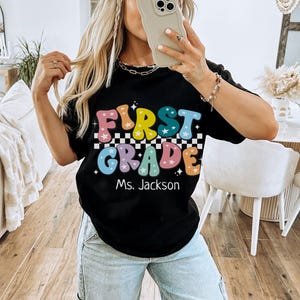 May include: Black t-shirt with the words "FIRST GRADE" in colorful, retro-style lettering, with a checkered pattern and stars. The name "Ms. Jackson" is below the text. The word "BLACK" is at the bottom.