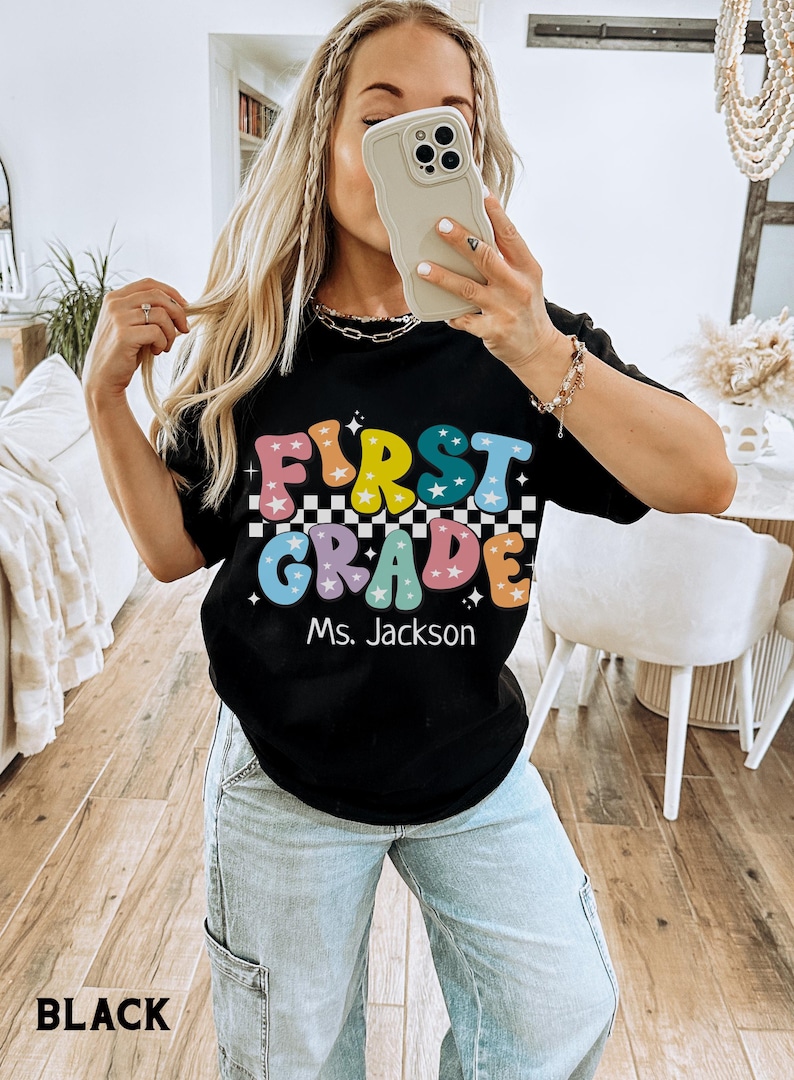 May include: Black t-shirt with the words "FIRST GRADE" in colorful, retro-style lettering, with a checkered pattern and stars. The name "Ms. Jackson" is below the text. The word "BLACK" is at the bottom.