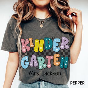 May include: A dark gray t-shirt with the words "KINDER GARDEN" in colorful, bubble letters with star accents. Below the text is "Mrs. Jackson". The shirt has a retro, vintage style.