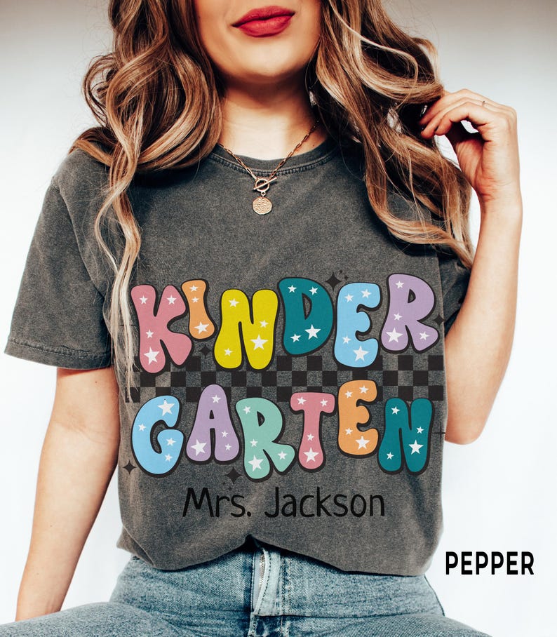 May include: A dark gray t-shirt with the words "KINDER GARDEN" in colorful, bubble letters with star accents. Below the text is "Mrs. Jackson". The shirt has a retro, vintage style.