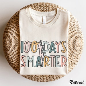 Pode incluir: Uma camiseta bege com o texto "100 Days Smarter" em letras coloridas e contornadas. O texto &eacute; estilizado com um gr&aacute;fico de raio.