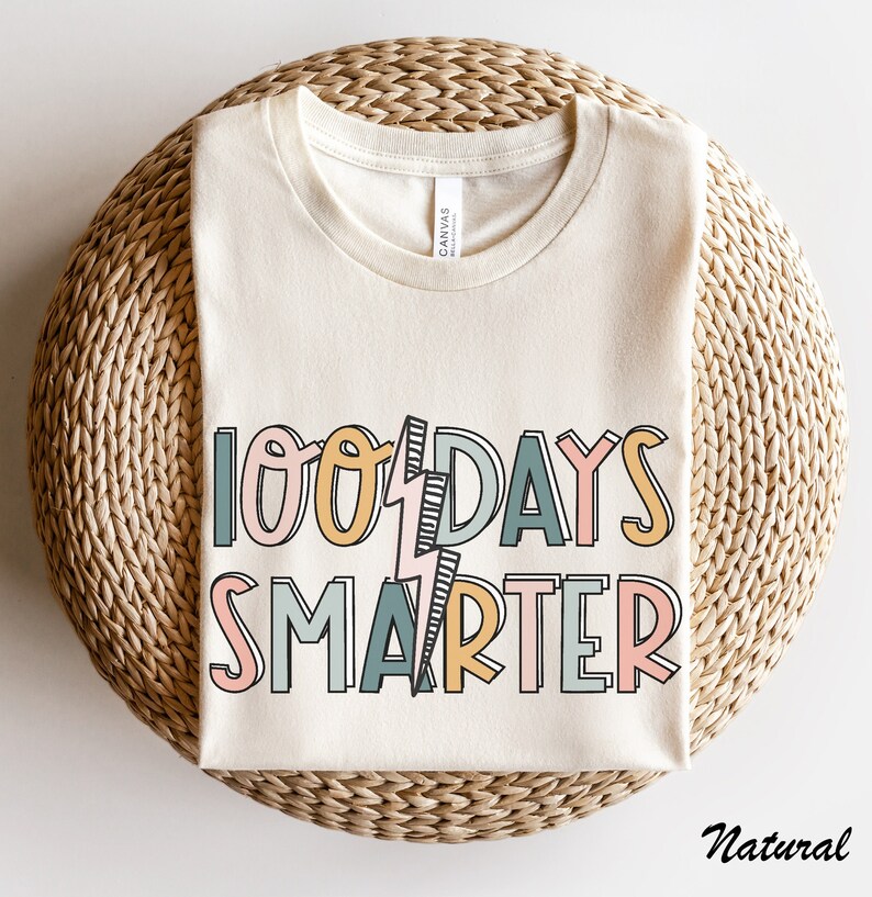 Pode incluir: Uma camiseta bege com o texto "100 Days Smarter" em letras coloridas e contornadas. O texto &eacute; estilizado com um gr&aacute;fico de raio.