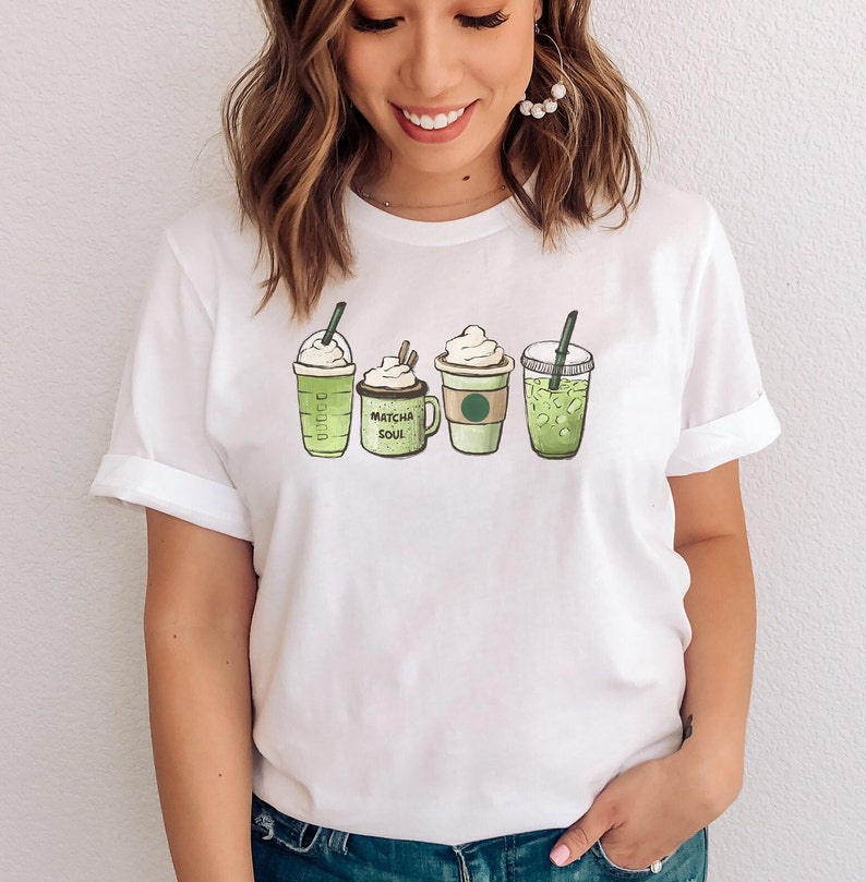 May include: White t-shirt with a graphic of four green matcha drinks. The drinks are a tall iced matcha latte, a matcha latte with whipped cream, a matcha latte with whipped cream and a straw, and a tall iced matcha latte with a straw. The text "Matcha Soul" is printed on the t-shirt.