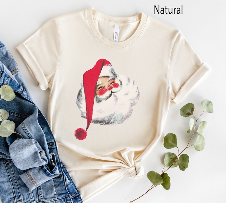 May include: A cream colored t-shirt with a vintage illustration of Santa Claus's face. The illustration is in red, white, and black.