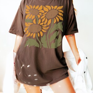 May include: A brown oversized t-shirt with a graphic print of three sunflowers in shades of yellow, orange, and brown. The sunflowers are stylized and have a vintage look.