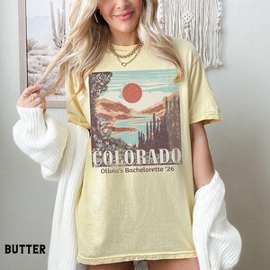 May include: Butter-colored t-shirt featuring a Colorado landscape design with a red sun, mountains, and a lake. The word "COLORADO" is printed in a bold font, with "Olivia's Bachelorette '26" below. The shirt is a relaxed fit.