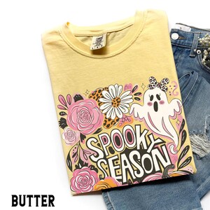 May include: A yellow t-shirt with a "Spooky Season" graphic featuring a ghost, flowers, and pumpkins. The shirt is folded and displayed with denim shorts. The word "BUTTER" is printed at the bottom of the image.