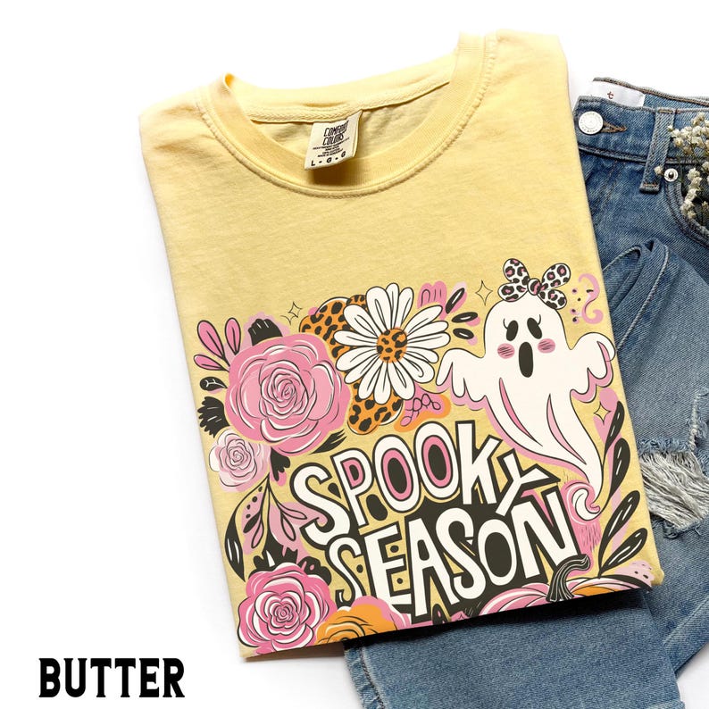 May include: A yellow t-shirt with a "Spooky Season" graphic featuring a ghost, flowers, and pumpkins. The shirt is folded and displayed with denim shorts. The word "BUTTER" is printed at the bottom of the image.