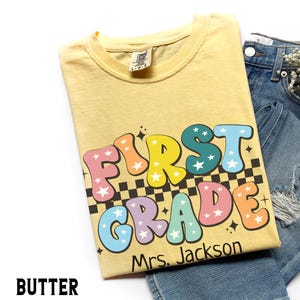 May include: A light yellow t-shirt with the words "FIRST GRADE" in colorful bubble letters with star accents. The shirt also includes the name "Mrs. Jackson" and a black and white checkered pattern.