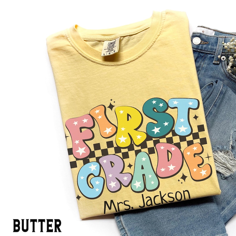 May include: A light yellow t-shirt with the words "FIRST GRADE" in colorful bubble letters with star accents. The shirt also includes the name "Mrs. Jackson" and a black and white checkered pattern.