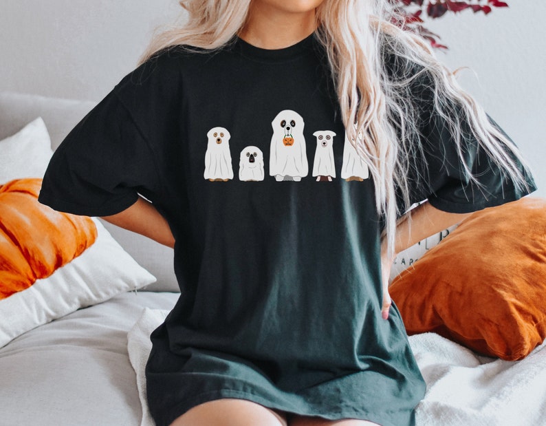 May include: A black t-shirt with a graphic of four dogs dressed as ghosts for Halloween. The ghosts are white and have black eyes. One ghost is holding a small orange pumpkin.