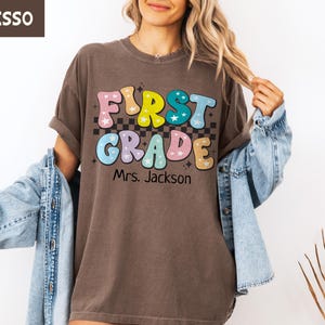 May include: Espresso-colored t-shirt with a colorful "First Grade" design, featuring stars and a checkered background. The shirt includes the name "Mrs. Jackson" below the text, perfect for teachers or students.