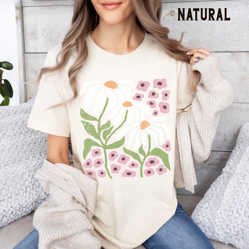 K&ouml;nnte beinhalten: Ein cremefarbenes T-Shirt mit einem grafischen Druck von wei&szlig;en G&auml;nsebl&uuml;mchen und rosa Blumen. Der Text "NATURAL" ist in der oberen rechten Ecke des Bildes sichtbar.