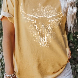 K&ouml;nnte beinhalten: Ein senfgelbes T-Shirt mit einem wei&szlig;en Aufdruck eines Stierkopfes mit floralen Designs. Der Text "Senf" ist am unteren Rand des Bildes sichtbar.