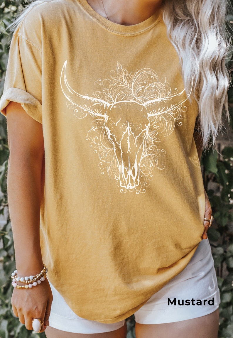 K&ouml;nnte beinhalten: Ein senfgelbes T-Shirt mit einem wei&szlig;en Aufdruck eines Stierkopfes mit floralen Designs. Der Text "Senf" ist am unteren Rand des Bildes sichtbar.
