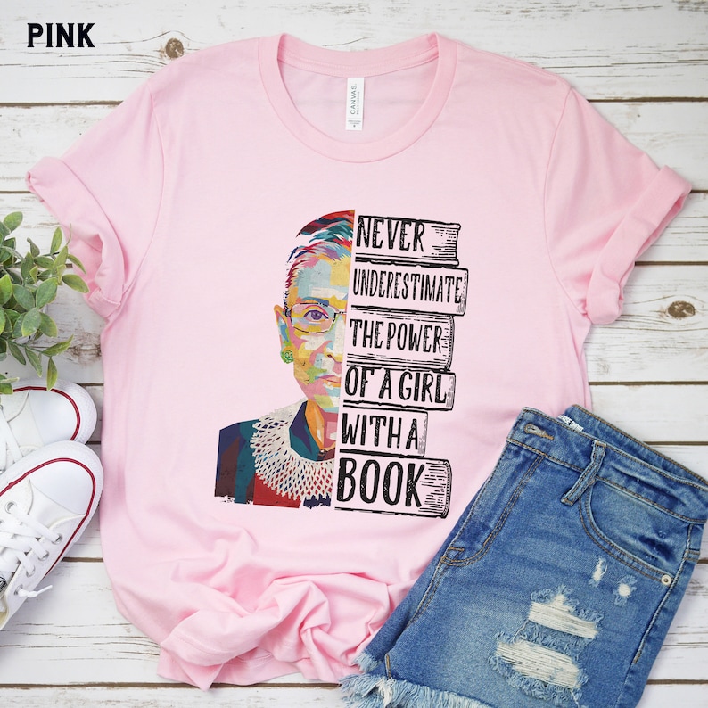 Op de afbeelding: Zachtroze T-shirt met een kleurrijk portret van een vrouw en de tekst "Never Underestimate The Power Of A Girl With A Book". Het shirt wordt gestyled met een spijkerbroekshort en witte sneakers.
