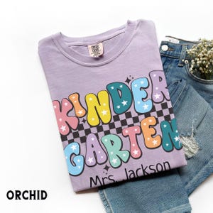 May include: Orchid-colored t-shirt with the words "KINDERGARTEN" in colorful bubble letters with star accents. The shirt includes the name "Mrs. Jackson" and is paired with blue jeans.