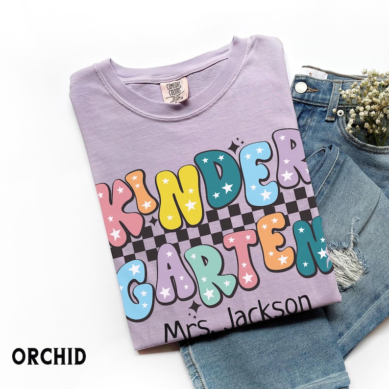 May include: Orchid-colored t-shirt with the words "KINDERGARTEN" in colorful bubble letters with star accents. The shirt includes the name "Mrs. Jackson" and is paired with blue jeans.