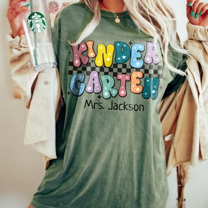 May include: Olive green t-shirt with the words "KINDER GARDEN" in colorful bubble letters, with stars and a checkered background. The name "Mrs. Jackson" is printed below. A clear Starbucks tumbler with a green straw is held in the left hand.