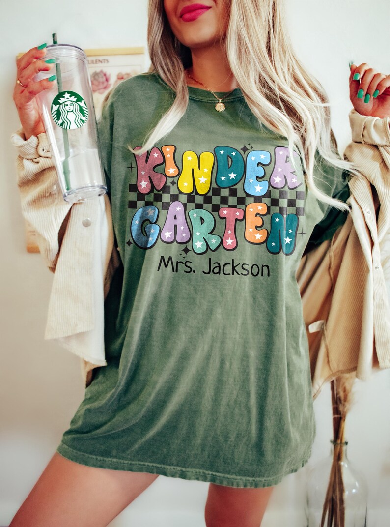 May include: Olive green t-shirt with the words "KINDER GARDEN" in colorful bubble letters, with stars and a checkered background. The name "Mrs. Jackson" is printed below. A clear Starbucks tumbler with a green straw is held in the left hand.