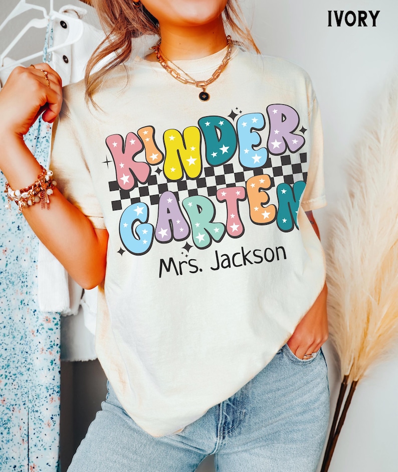 May include: Ivory-colored t-shirt with the words "KINDER GARDEN Mrs. Jackson" in colorful, retro-style lettering. The word "KINDER" is in yellow, blue, pink, and green. The word "GARDEN" is in blue, purple, orange, and green. The shirt has a checkered pattern.