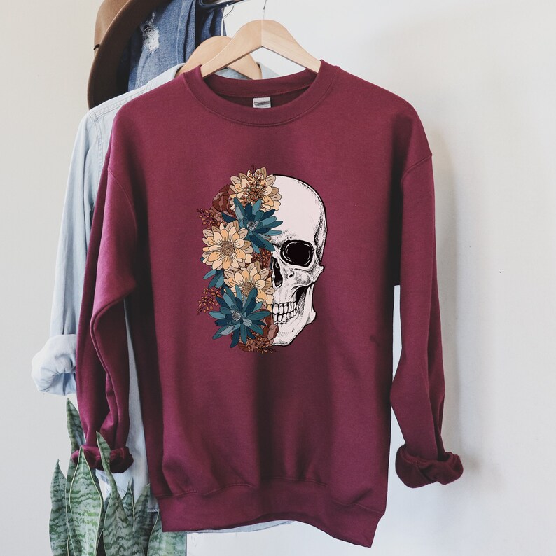 May include: A maroon sweatshirt with a white skull design. The skull is partially covered with colorful flowers in shades of blue, yellow, and brown.