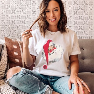May include: A woman wearing a white t-shirt with a vintage illustration of Santa Claus. The shirt has a red and white Santa hat and a white beard. She is wearing blue jeans with rips at the knees and is sitting on a couch.