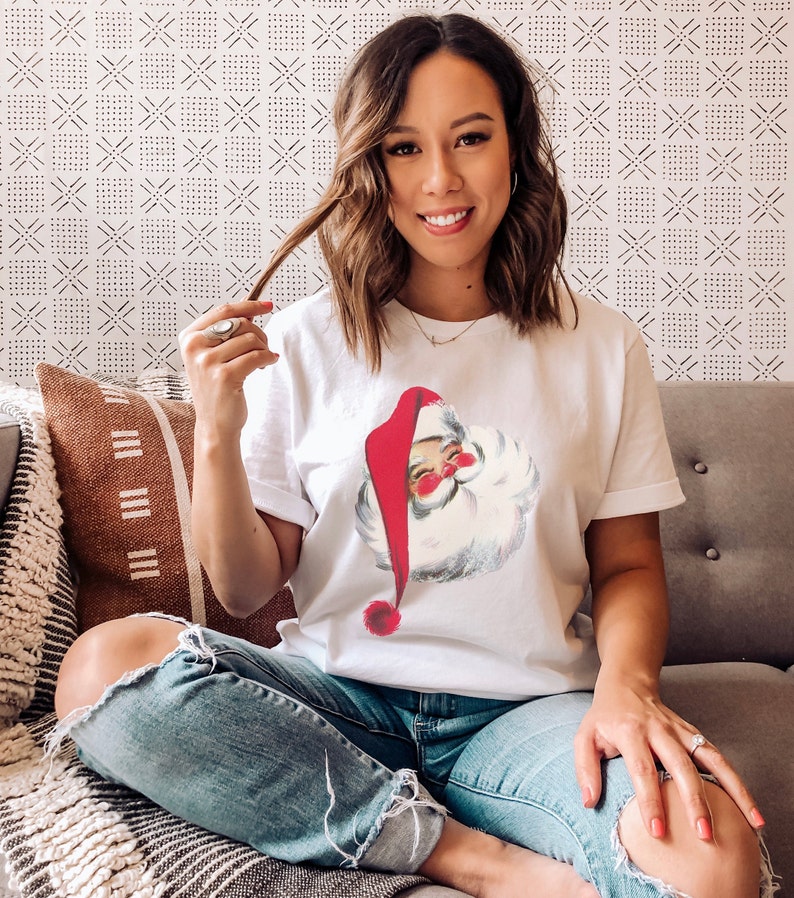 May include: A woman wearing a white t-shirt with a vintage illustration of Santa Claus. The shirt has a red and white Santa hat and a white beard. She is wearing blue jeans with rips at the knees and is sitting on a couch.