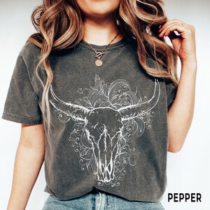 K&ouml;nnte beinhalten: Ein dunkelgraues T-Shirt mit einem wei&szlig;en Stierkopf-Motiv und einem floralen Design. Der Text "PEPPER" ist unten rechts auf dem Shirt aufgedruckt.
