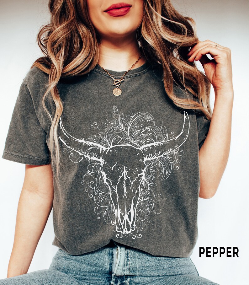 K&ouml;nnte beinhalten: Ein dunkelgraues T-Shirt mit einem wei&szlig;en Stierkopf-Motiv und einem floralen Design. Der Text "PEPPER" ist unten rechts auf dem Shirt aufgedruckt.