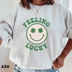 May include: A gray sweatshirt with the text "Feeling Lucky" in green and white lettering. A green smiley face with shamrocks for eyes is centered on the sweatshirt.