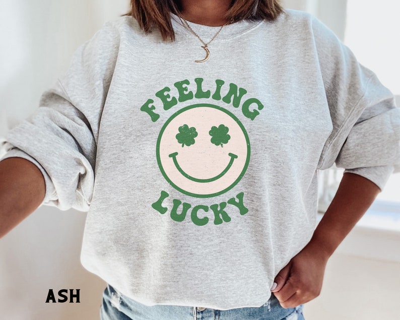 May include: A gray sweatshirt with the text "Feeling Lucky" in green and white lettering. A green smiley face with shamrocks for eyes is centered on the sweatshirt.