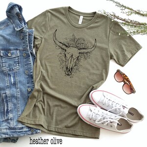 Peut inclure: Un t-shirt vert olive avec un motif noir de cr&acirc;ne de taureau avec des accents floraux.