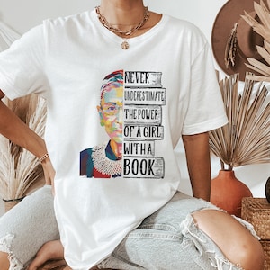 Op de afbeelding: Wit T-shirt met een kleurrijke afbeelding van een vrouwengezicht en de tekst "NEVER UNDERESTIMATE THE POWER OF A GIRL WITH A BOOK". Het ontwerp is verdeeld tussen het gezicht en een stapel boeken. Het shirt heeft korte mouwen.