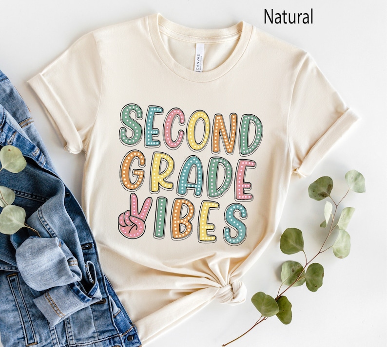 May include: A beige t-shirt with the text "Second Grade Vibes" in colorful letters with polka dots. The text is in a retro style font.