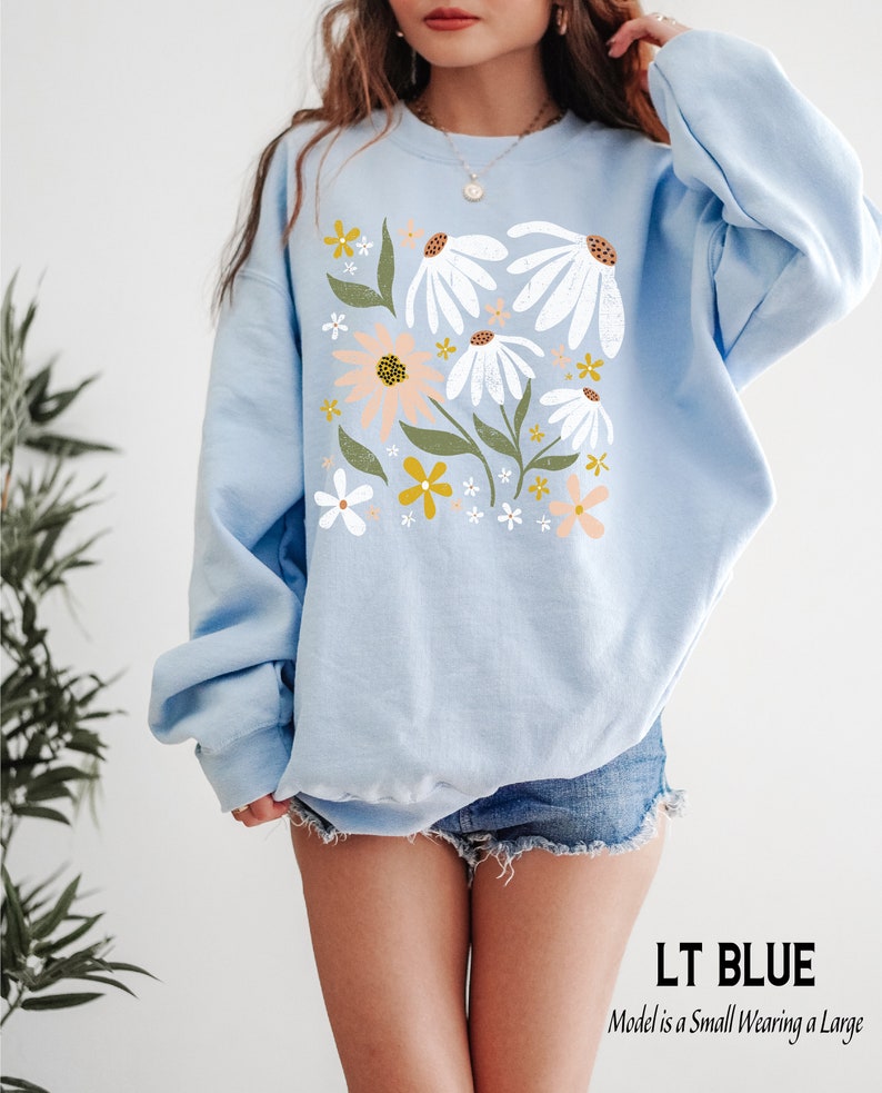 K&ouml;nnte beinhalten: Ein hellblauer Pullover mit einem Blumendruck mit wei&szlig;en, rosa und gelben Blumen. Das Model tr&auml;gt Gr&ouml;&szlig;e L.