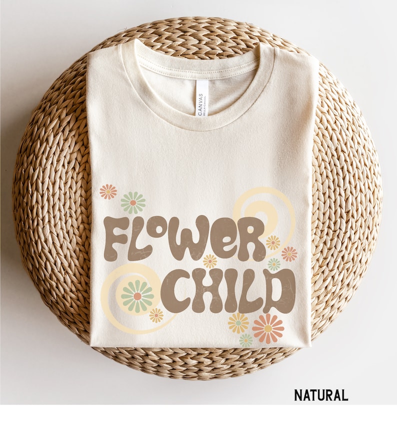 Puede incluir: Una camiseta doblada, de color crema, con las palabras "FLOWER CHILD" en letras marrones de estilo retro. El dise&ntilde;o incluye flores y espirales de colores pastel. La camiseta est&aacute; sobre una superficie circular tejida. La palabra "NATURAL" est&aacute; impresa en la parte inferior.
