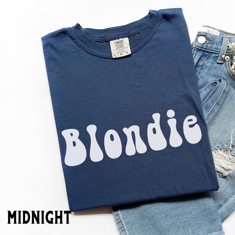 Puede incluir: Una camiseta azul marino con la palabra "Blondie" en blanco, con letras de estilo retro. La camiseta est&aacute; doblada y colocada junto a unos pantalones cortos vaqueros. La palabra "MIDNIGHT" est&aacute; impresa en negro en la esquina inferior izquierda.