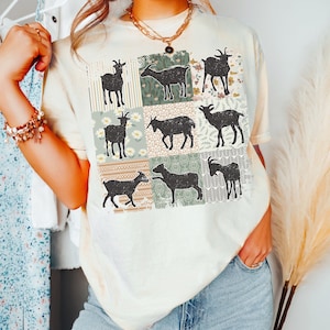 May include: A cream-colored t-shirt with a patchwork design featuring silhouettes of goats in various poses. The design is made up of squares with different patterns, including stripes, floral prints, and geometric designs.