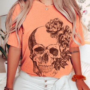 May include: Terracotta-colored t-shirt featuring a detailed skull graphic with floral accents. The shirt has short sleeves and is paired with white pants. The word "TERRACOTTA" is printed at the bottom.