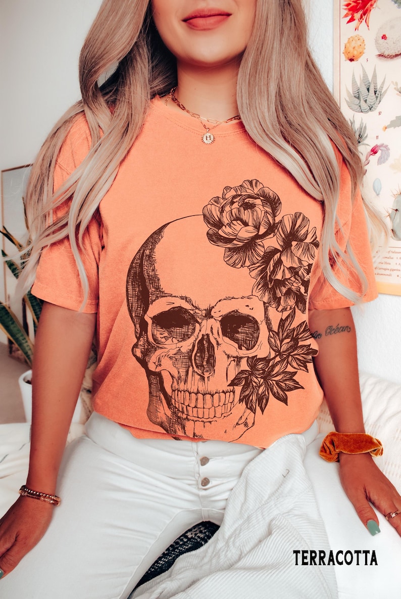 May include: Terracotta-colored t-shirt featuring a detailed skull graphic with floral accents. The shirt has short sleeves and is paired with white pants. The word "TERRACOTTA" is printed at the bottom.