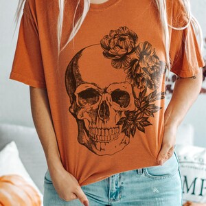 May include: Orange short-sleeved t-shirt featuring a black skull design with floral accents. The word "YAM" is printed in the upper left corner. The model is wearing light blue jeans.