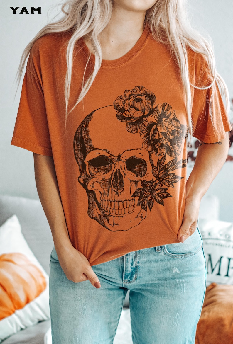 May include: Orange short-sleeved t-shirt featuring a black skull design with floral accents. The word "YAM" is printed in the upper left corner. The model is wearing light blue jeans.