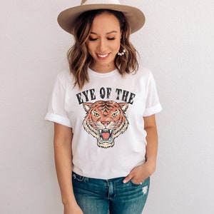 May include: White t-shirt with a vintage-style tiger graphic and the text "Eye of the Tiger". The model is wearing blue jeans and a brown fedora.