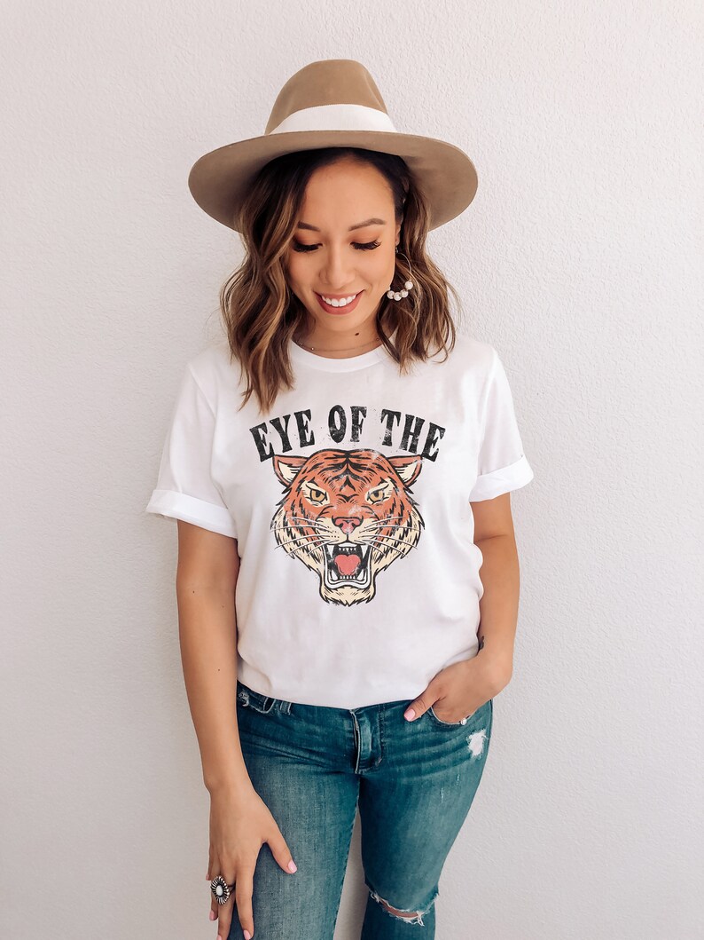 May include: White t-shirt with a vintage-style tiger graphic and the text "Eye of the Tiger". The model is wearing blue jeans and a brown fedora.