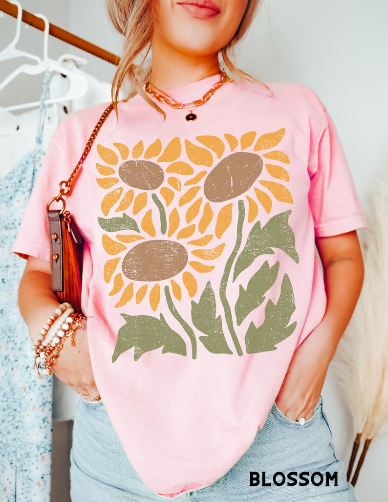 May include: A pink t-shirt with a graphic of three stylized sunflowers in shades of yellow, brown, and green.