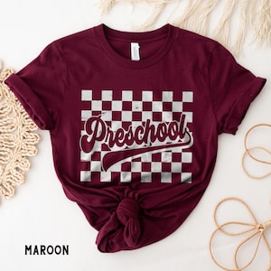 Pu&ograve; includere: T-shirt bordeaux con motivo a scacchi bianchi e grigi e la scritta "Preschool" in un font retr&ograve;. La maglietta &egrave; annodata in basso. La parola "MAROON" &egrave; stampata in basso a sinistra.